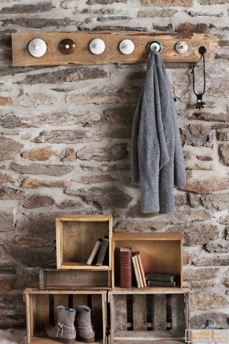 Guardaroba rustico fai-da-te in camera con muro in pietra; pioli di cappotto fatti da vecchi coperchi di porcellana e vecchie casse di legno sul pavimento come deposito